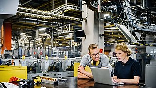Zwei Personen sitzen vor einem Laptop in einer Fabrikhalle. Drumherum sind industrielle Anlagen.